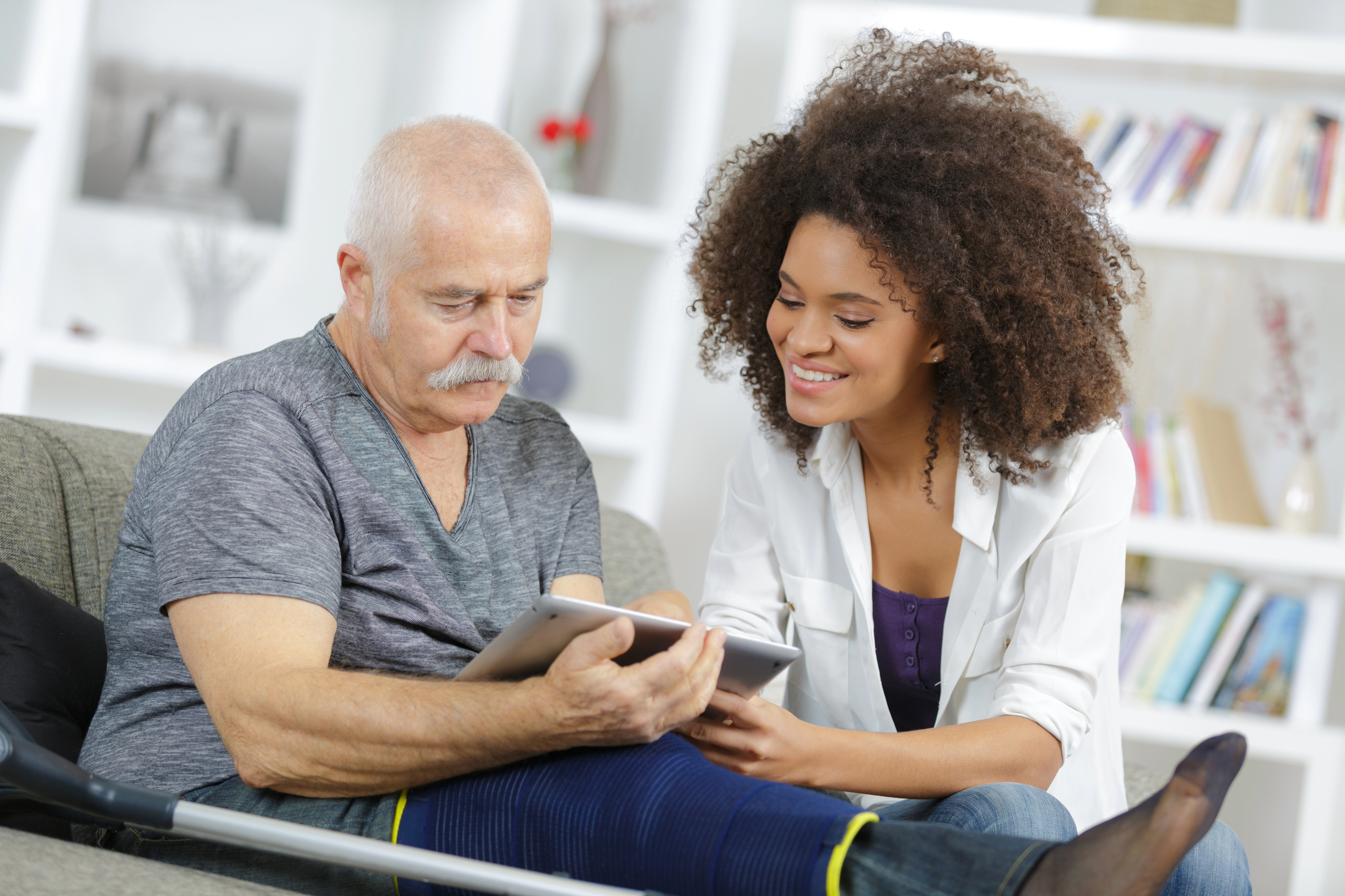 A home health aide and an elderly client sitting on a couch looking at a tablet