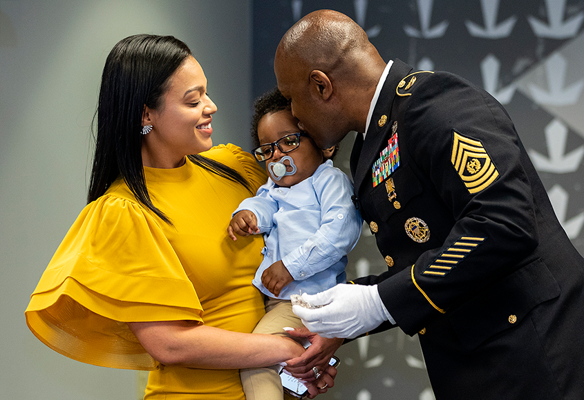 Baby held by woman in yellow dress and being kissed by veteran. 