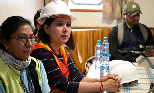 Two construction workers in safety gear sitting at a table during a meeting, with another person in casual wear looking at a phone in the background.