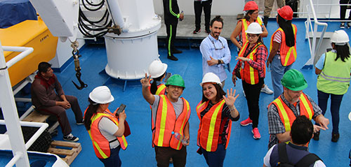 Group of workers in safety gear including helmets and reflective vests on a ship, some waving at the camera.