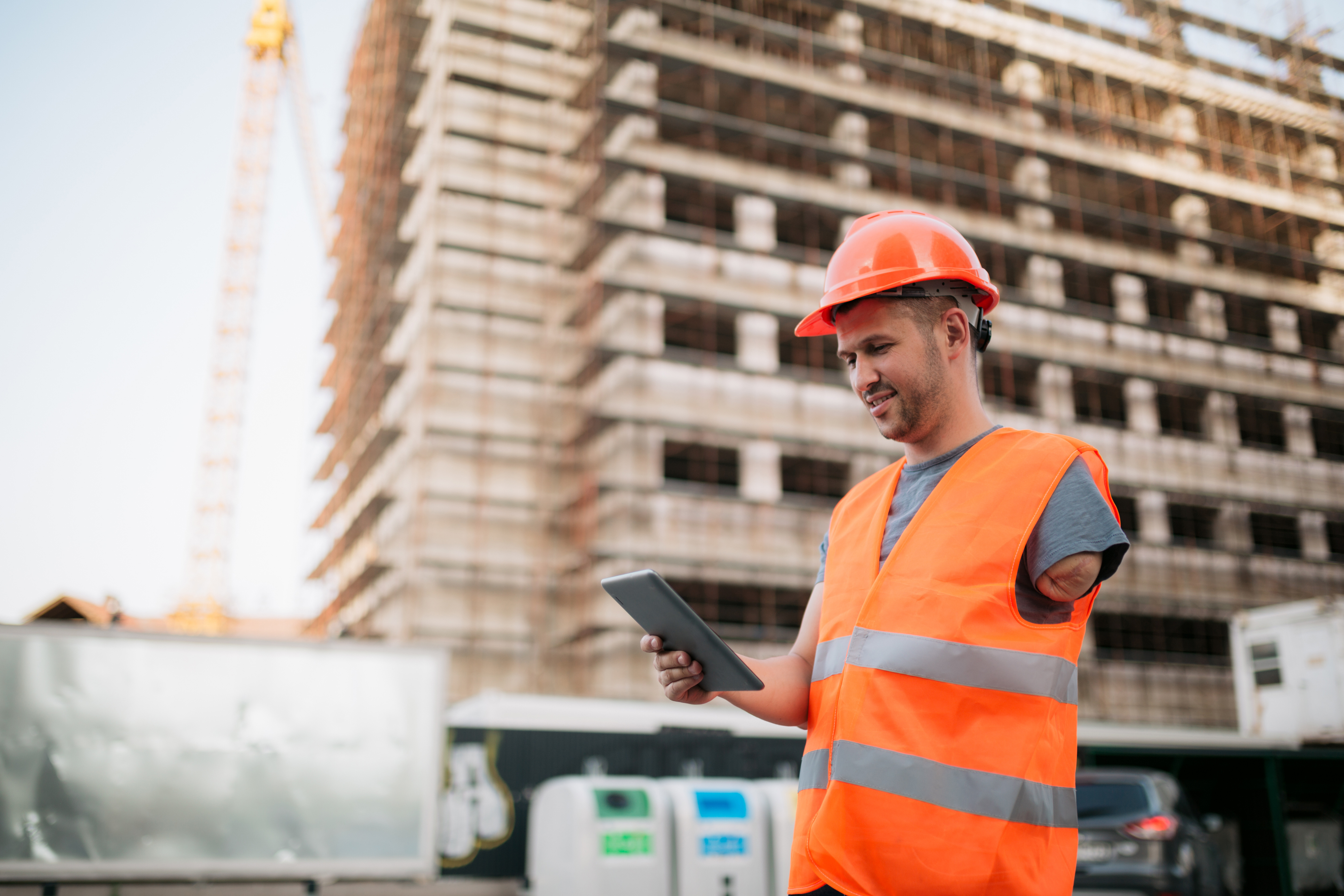 A construction worker with a limb difference in an orange hard hat and vest reads from a tablet in front of a construction site.