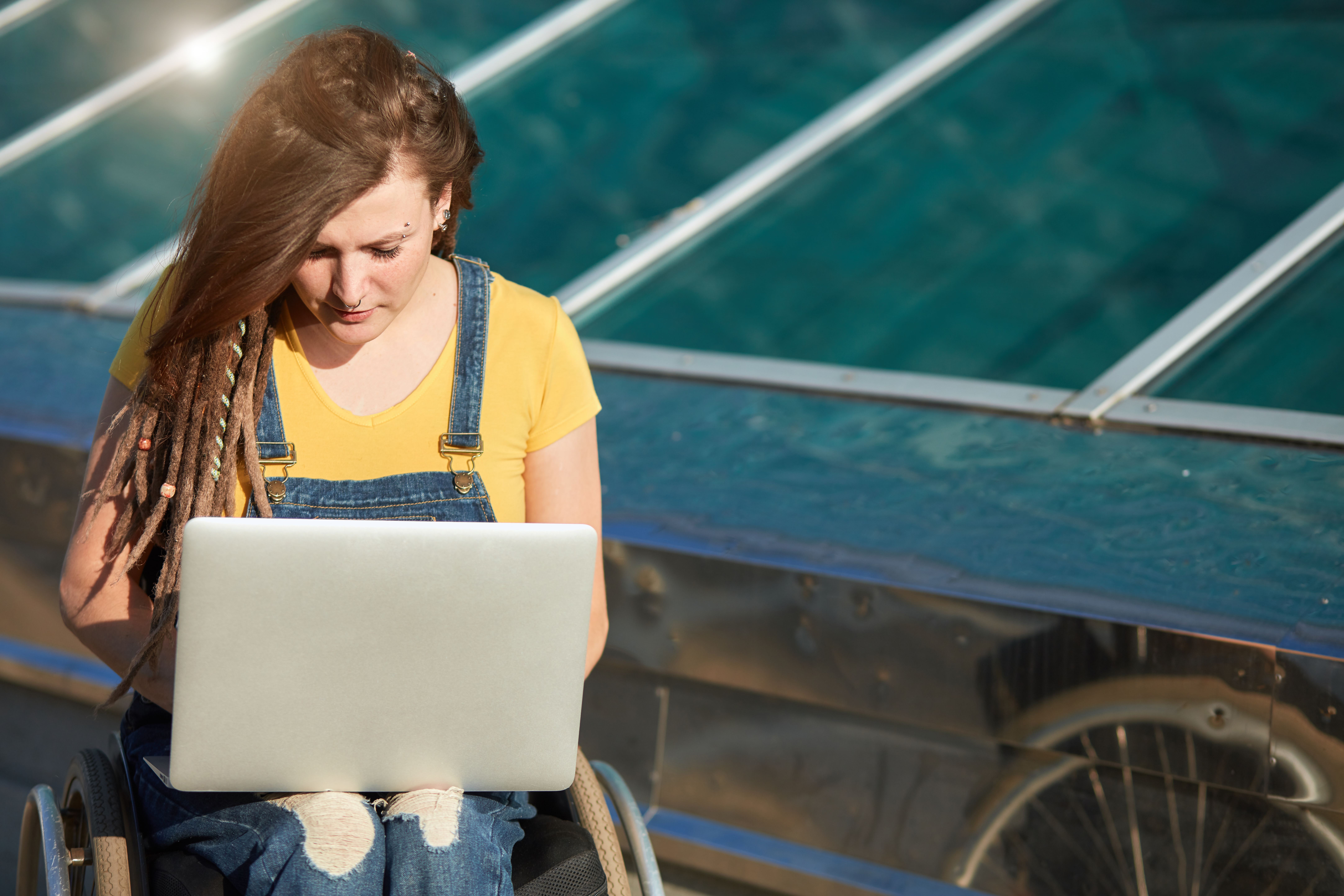 A wheelchair user wearing a yellow shirt and overalls types on a laptop in front of a wall of solar panels.