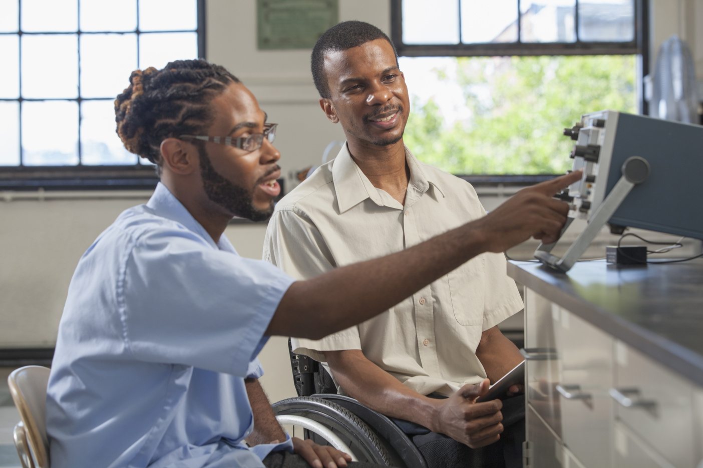 Two people, one with cornrows in their hair and a beard, and the other using a wheelchair, sit in a laboratory environment looking at a microscope, with a window open in the background.