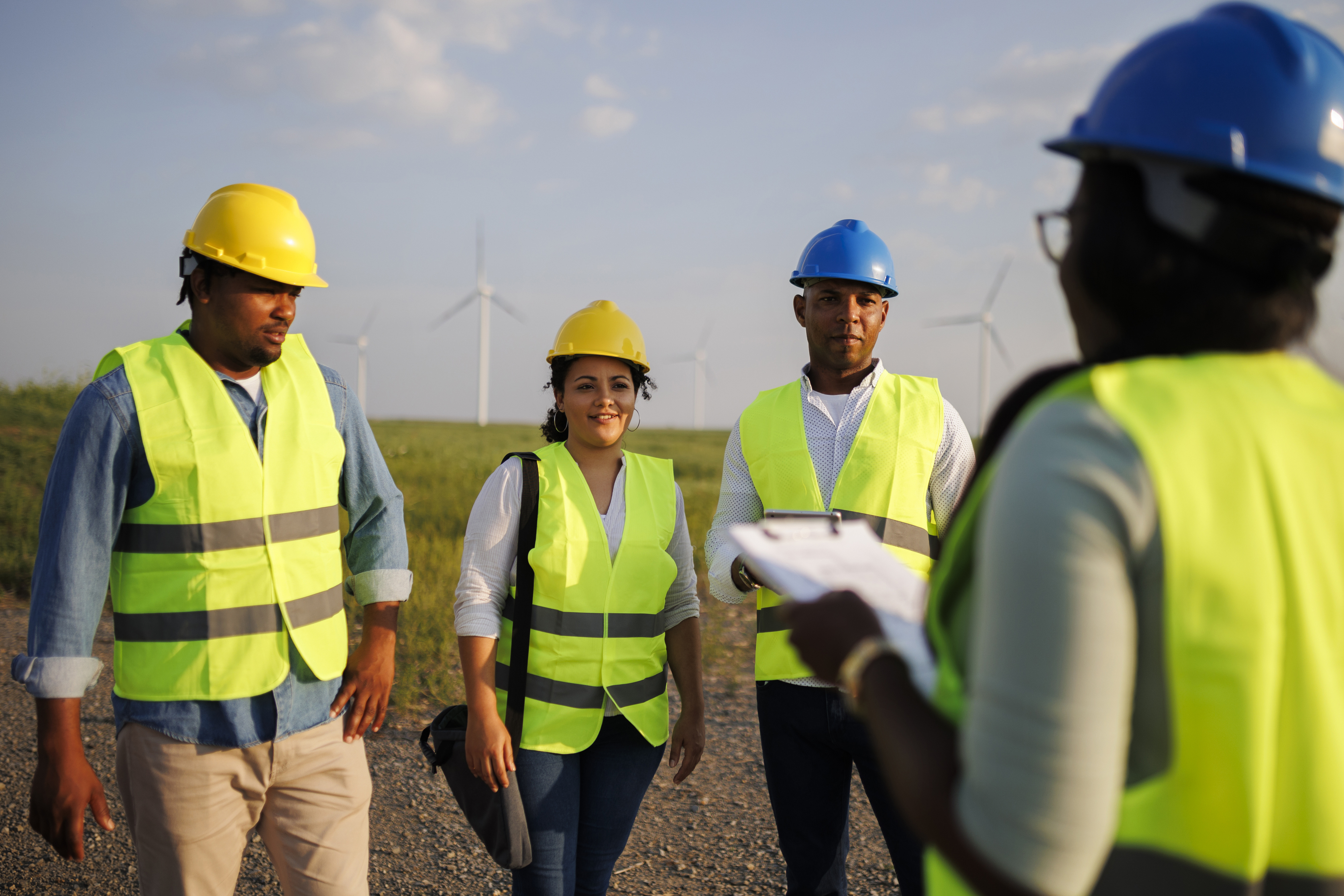 Workers wearing blue and yellow helmets and safety vests stand in a field, facing another worker wearing similar equipment and holding a clipboard. Electric power windmills are visible in the background.