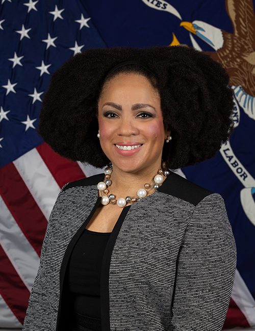 Black woman with shoulder length natural hair smiling at the camera in front of the American and U.S. Department of Labor flags. 