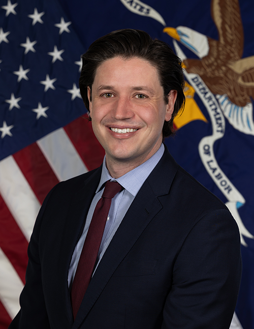 A white man with medium length brown hair wearing a blue suit in front of an American flag backdrop