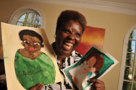 Lois Curtis smiling wearing a black and white patterned top holding two of her paintings. One is a painting of Lois herself and one is of her friend Elaine Wilson who joined the Olmstead case with her.