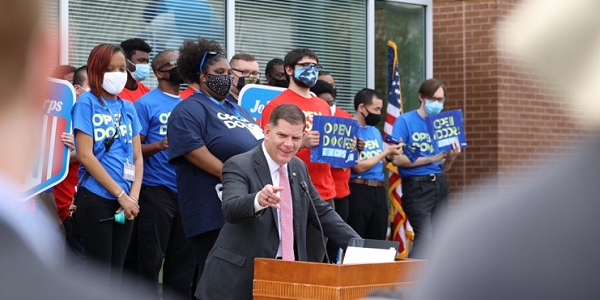 Secretary Walsh speaks at a podium. Behind him stand Job Corps students wearing shirts that say 'Open Doors.' 