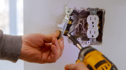 Close-up shot of an electrical outlet mid-installation. One hand holds a light switch and the other holds a drill. 