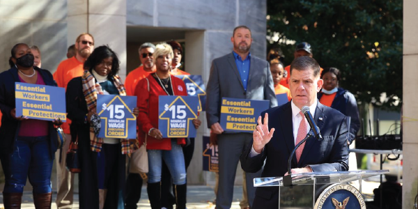 Secretary Walsh speaks at a podium with the department's seal in front of a crowd holding signs saying '$15 minimum wage Executive Order.' 