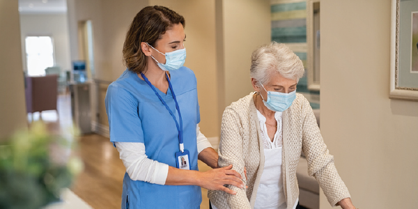 A caregiver in scrubs and face mask helps an older woman in face mask walk down a hallway. 