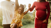 Two Black adults, one pregnant, hold a young childs hands so she can swing as they walk.