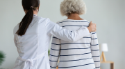 A woman in a medical coat helps an older woman walk.