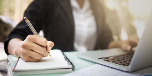 A young woman in professional clothes takes notes while sitting at a laptop.