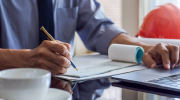 Close-up of a man signing a paycheck at a desk with blueprints and a hardhat in the background. 