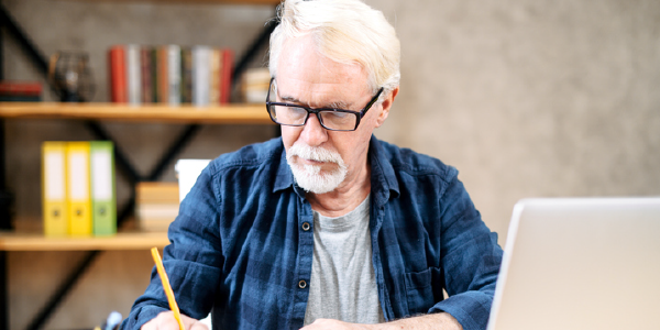 An older man in eyeglasses is using a laptop and writing in notebook. 