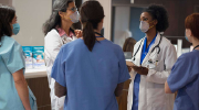 A group of medical professionals chat wearing scrubs and surgical masks. 