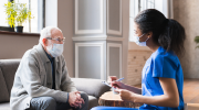 A healthcare worker in scrubs chats with an older patient at home.