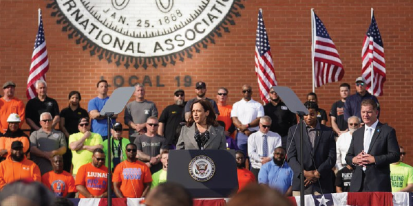 Vice President Kamala Harris speaks at a podium while Secretary Walsh stands to the side. Workers stand behind them.