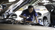 A woman in mechanics overalls leans under the raised hood of a car in a professional garage, examining the engine. 