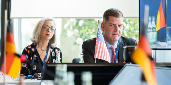 Secretary Walsh and ILAB head Thea Lee sit at a table behind an American flag. German flags are visible in the foreground. 