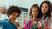 A teacher sits with two children in a classroom. Colored pencils sit in cups on the desk.