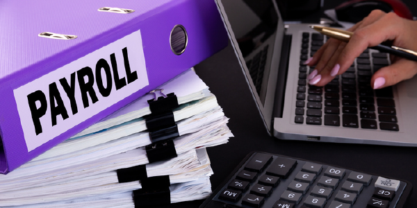 A womans hand hovers over a laptop keyboard. In the foreground are a calculator and a stack of papers below a binder labeled payroll.