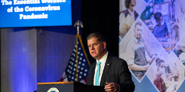 Secretary Walsh speaks at a podium in front of a collage of essential workers. 