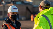 A man in a safety vest, mask and hardhat, holding a clipboard, chats with another man in a reflective jacket. 