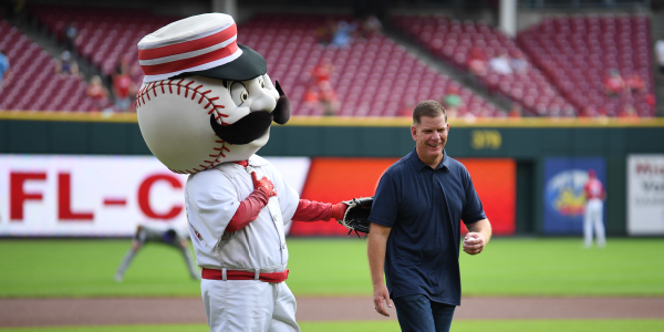 Secretary Walsh grins on a baseball field, while a mascot with the head of a baseball, sporting a cap and comically luxurious mustache, slaps him on the back. 