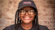 A young woman smiles at the camera. She has glasses and dreadlocks and wears a baseball cap.