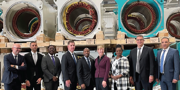 Secretary Walsh stands with eight men and women in professional clothing. Large machine parts are stacked behind them on pallets. 