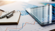 A pen on a notepad, a tablet whose screen shows charts, and print-outs of line graphs and bar graphs sit on a table. 