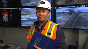 A man in a hardhat and reflective vest stands before a wall of screens showing images from mine sites.