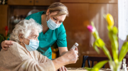 In a kitchen, a woman in scrubs and face mask helps an older woman, also wearing a face mask, to read a prescription bottle.