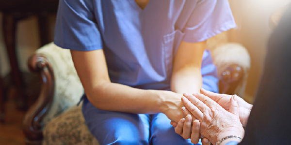 Close-up photo of a woman in blue scrubs in an armchair, holding the hands of an older woman.