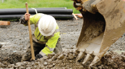 A man in hardhat and reflective vest digs a trench by the bucket of an excavator.