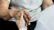 A close-up shot of an older woman taking the hand of a younger woman in a white medical coat.