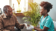 In a living room, a young woman in scrubs and medical gloves holds the hand of an older man. 