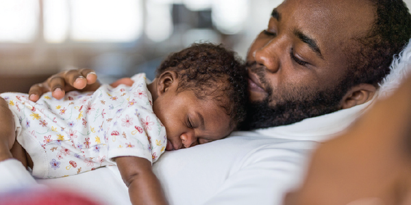 A father rests at home, holding his sleeping baby.