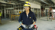 A woman in hard hat, toolbelt and safety goggles stands confidently in a building undergoing renovation.