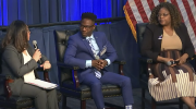 Three professionally dressed people sit on a stage, speaking in a panel.