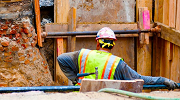 A worker wearing a safety vest and hard hat stands inside a trench that is properly shored up to prevent cave-ins.