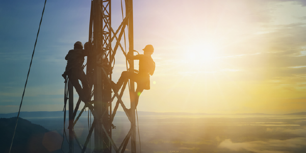 Telecom workers, silhouetted against the sun, scale on a telecommunications tower.