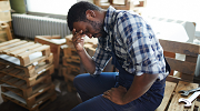 A worker with a plaid shirt sits in a room full of pallets and tools, holding his head in one hand. 