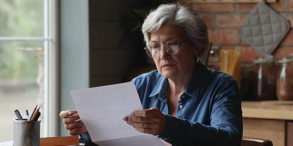 An older woman sits at the kitchen table, reading a document with a serious expression.