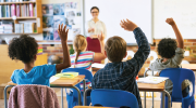 A teacher speaks as children, seated at their desks, raise their hands to join a classroom discussion.