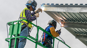 Two workers stand on an elevated platform to weld a metal roof. They are wearing fall protection harnesses, welding helmets, gloves and high visibility clothing.