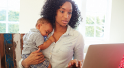 A woman holding an infant scrolls through a laptop.