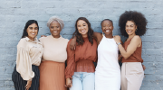 Five professionally dressed women of different racial backgrounds pose for a photo in front of a brick wall. 